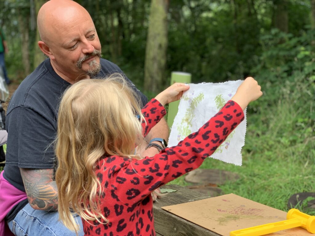 A little girl with blond hair and wearing a red leopard print top holds up a sheet of paper with some natural prints on it. A man smiles at it proudly. They are taking part in Washington Wetland Centre's Clay Faces and Natural Printing activity.