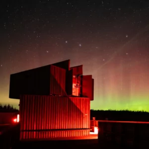 View of Kielder Observatory's wooden wall against a night sky backdrop. Green Northern Lights tinge the horizon and stars stud the pink sky.