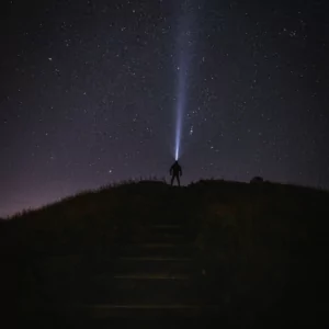The small silhouette of a man can be seen standing at the top of a hill. He wears a head torch and looks up at the constellations in the night sky, the beam of light shining upwards. Photo by Jim Scott.