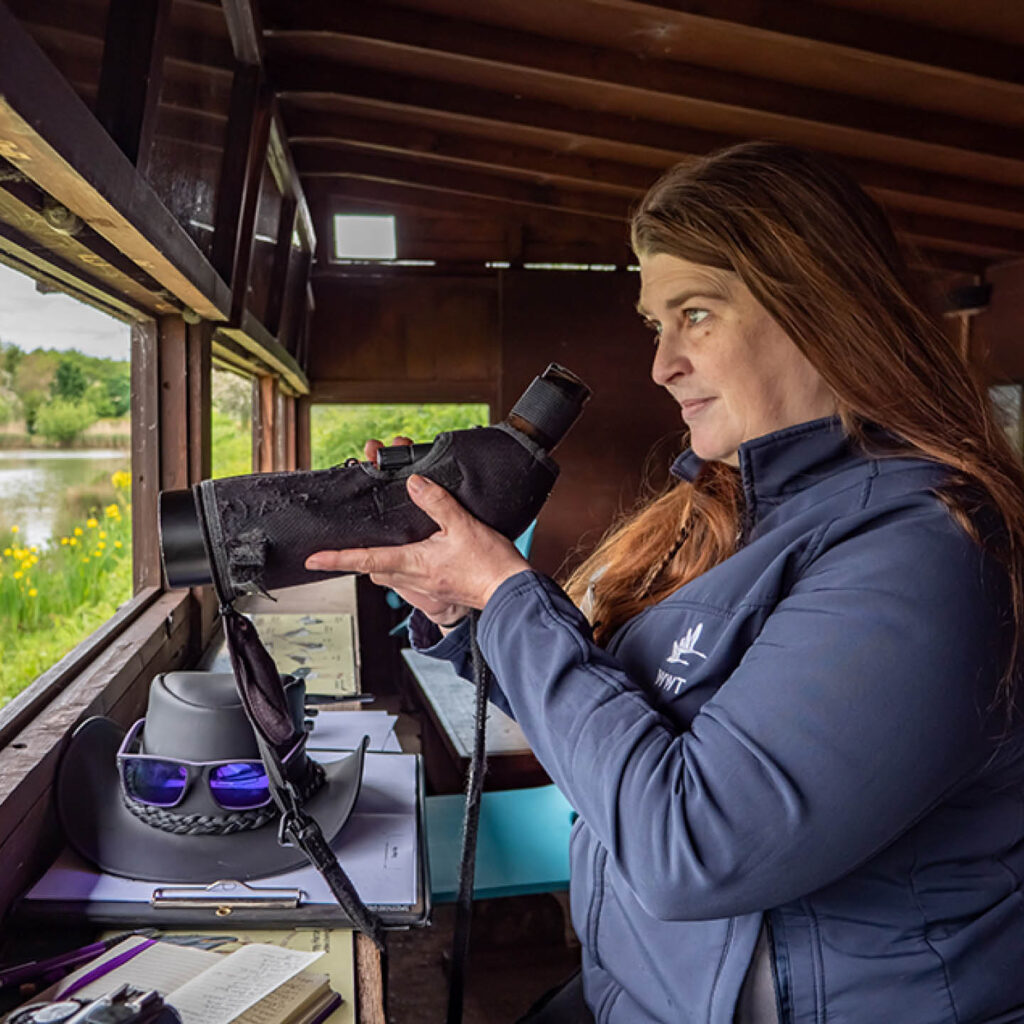 Wildlife volunteer Melissa is a white woman with long brown hair. She stands in a wooden hide at Washington Wetlands Centre. She holds a large telescope in her hands and looks out through the window, a smile on her face.