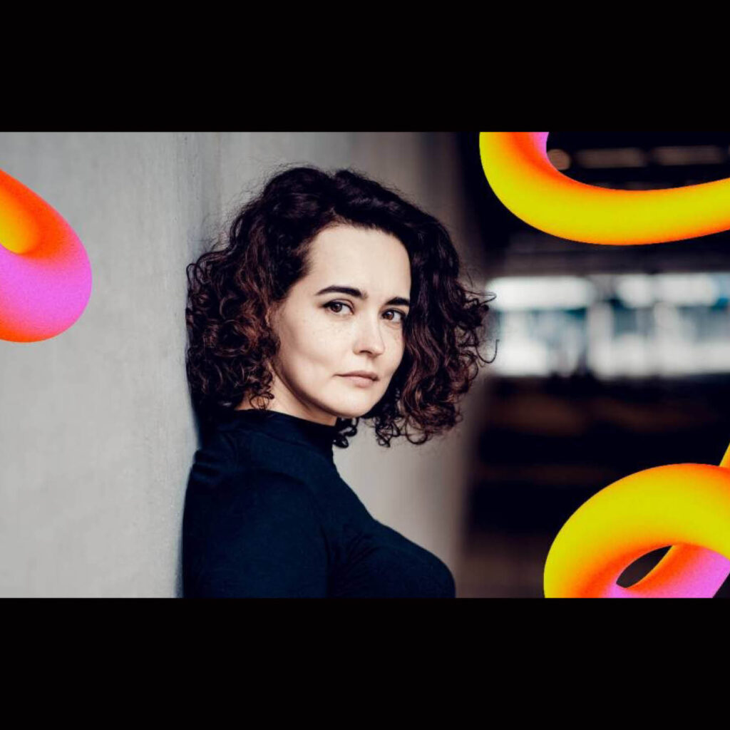 Violinist Alena Baeva is a white woman with black, shoulder length curly hair. She leans against a concrete wall and turns her head to look at the camera.
