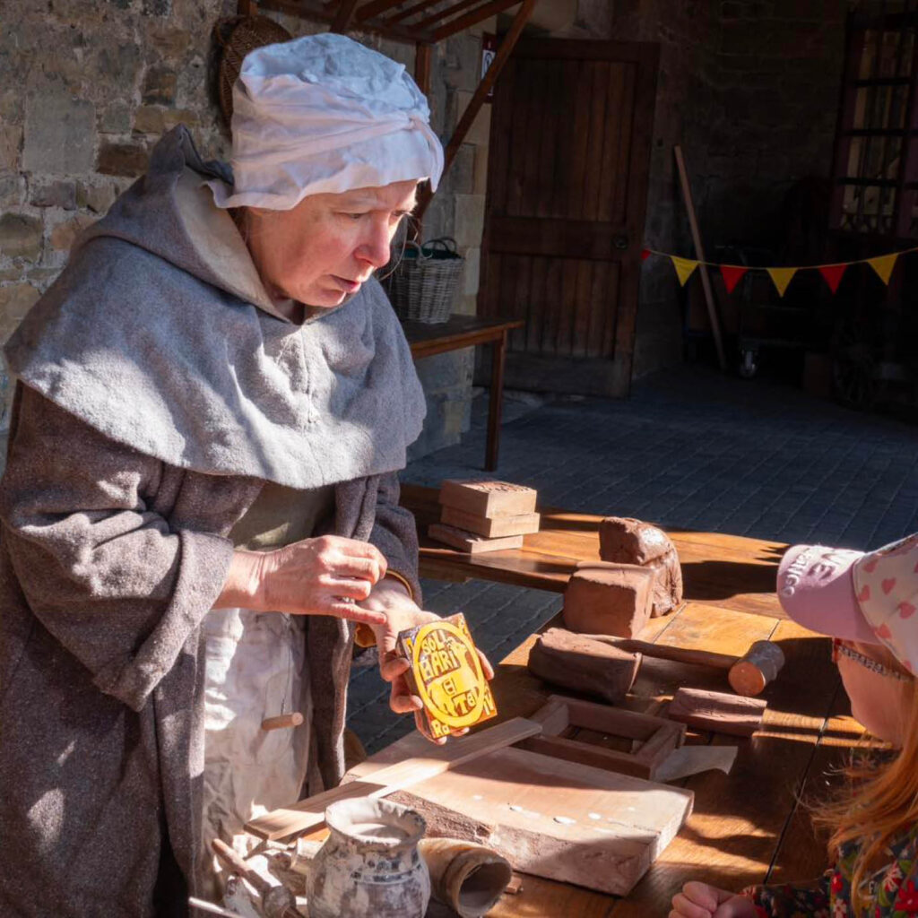 A woman dressed in a grey medieval style tunic and a white cloth hat holds out a tile stamp to show a little girl. She is one of the artisans at Alnwick Castle's Artisan Courtyard.