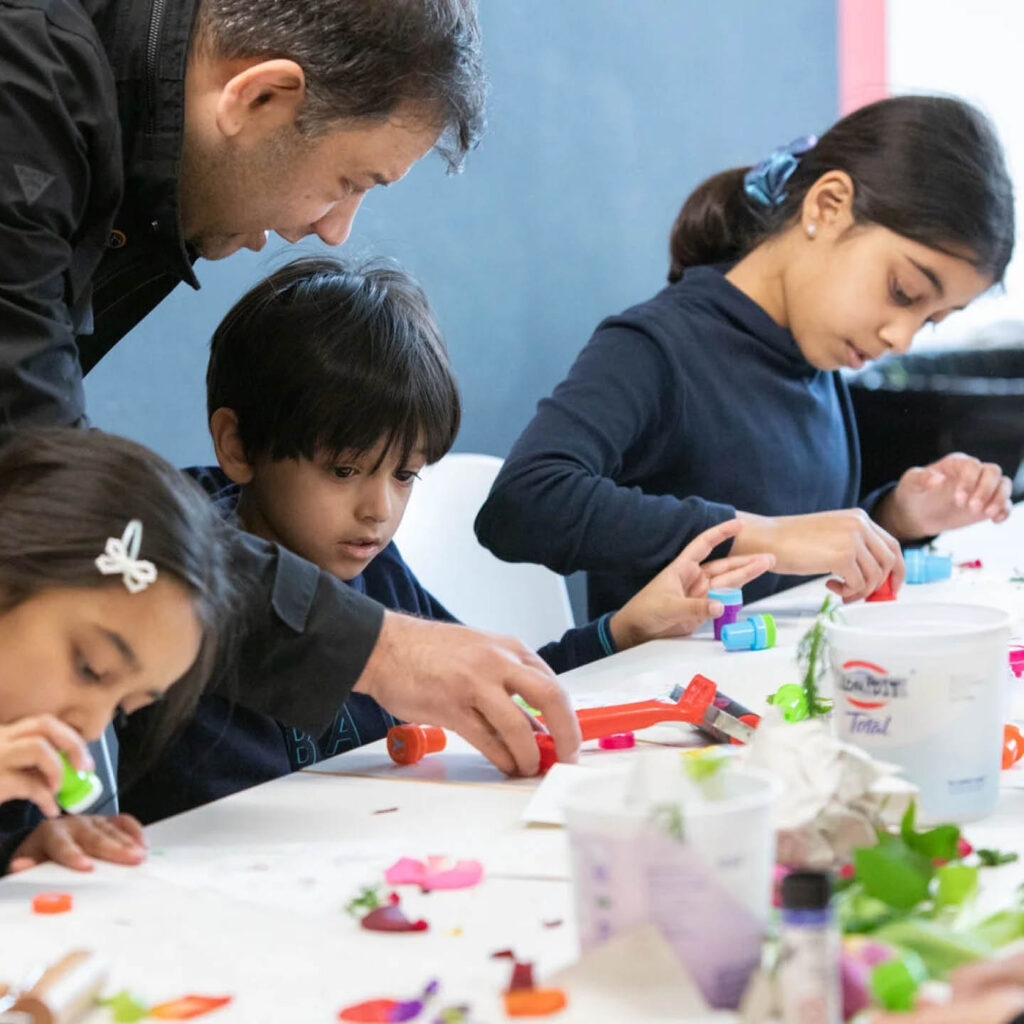 Three young children sit at a table. They are creating art with differently coloured stamps. A man leans over and helps the little boy with his work. They are all enjoying an activity at one of BALTIC's Big Days Out.