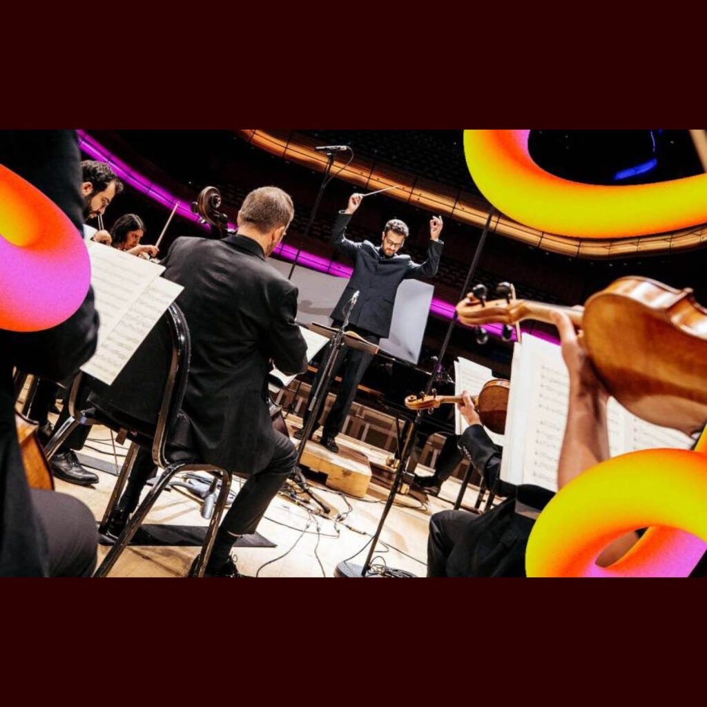 A conductor raises both arms in the air while looking at his sheet music. In the foreground, seated musicians play their instruments.