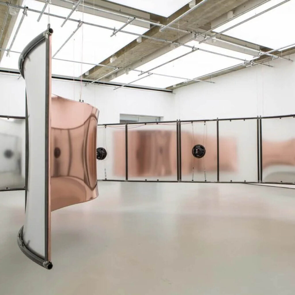 Large curved structures that look like metal walls fill a warehouse space. They have a coppery finish to them. Some have electric elements attached the their centres. This is a piece artist Hannah Perry called GUSH.