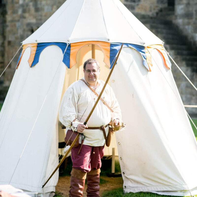 Andy Rice, a master archer at Alnwick Castle, wears a medieval style white tunic and red trousers with brown boots. He carries a long bow in one hand. Behind him is a small white tent.