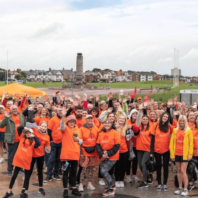 A huge crowd of people gather at Spanish City in Whitley Bay. They are all dressed in orange t-shirts with Maggie's across the front. They raise their hands and cheer at the camera.