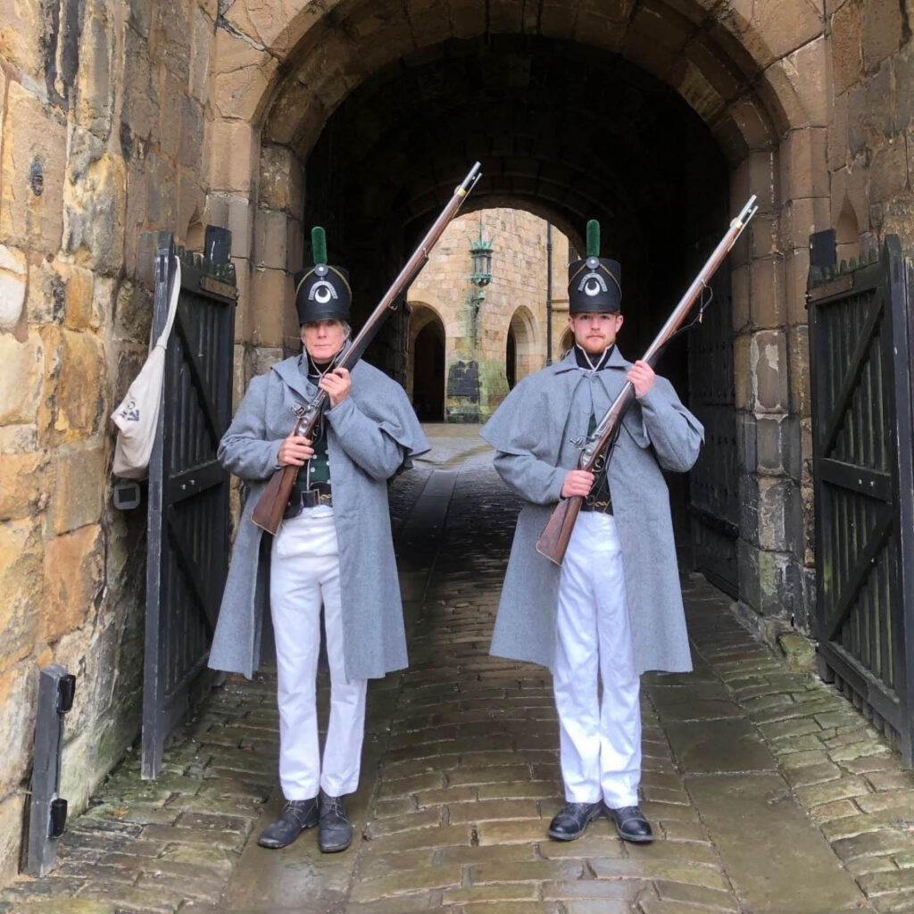Two men stand in the stone archway to Alnwick Castle. They are dressed in grey peacoats, white trousers and tall black hats. They both hold a musket across their bodies. They are part of the Percy Tenantry Volunteer Army.