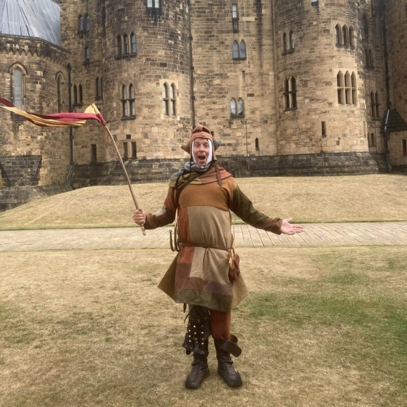 A man dressed in a brown patchwork jesters outfits stands on the lawn outside Alnwick Castle. He waves a red flag in one hand and gestures widely with his other. He opens his mouth in merriment.