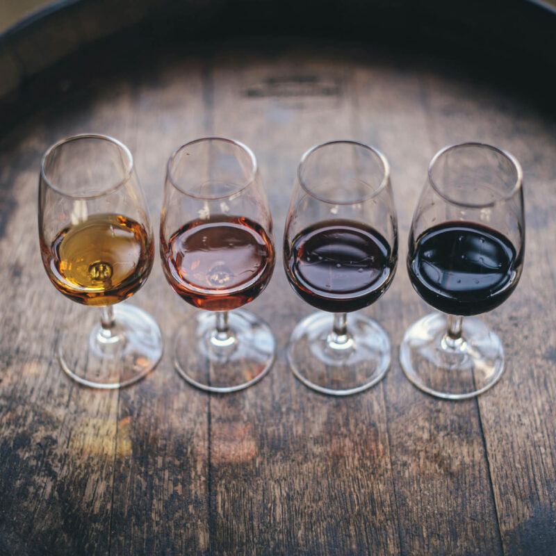 Four wine glasses are lined up on a wooden barrel table. Each is filled with a small amount of different coloured wine, from white to deep red.