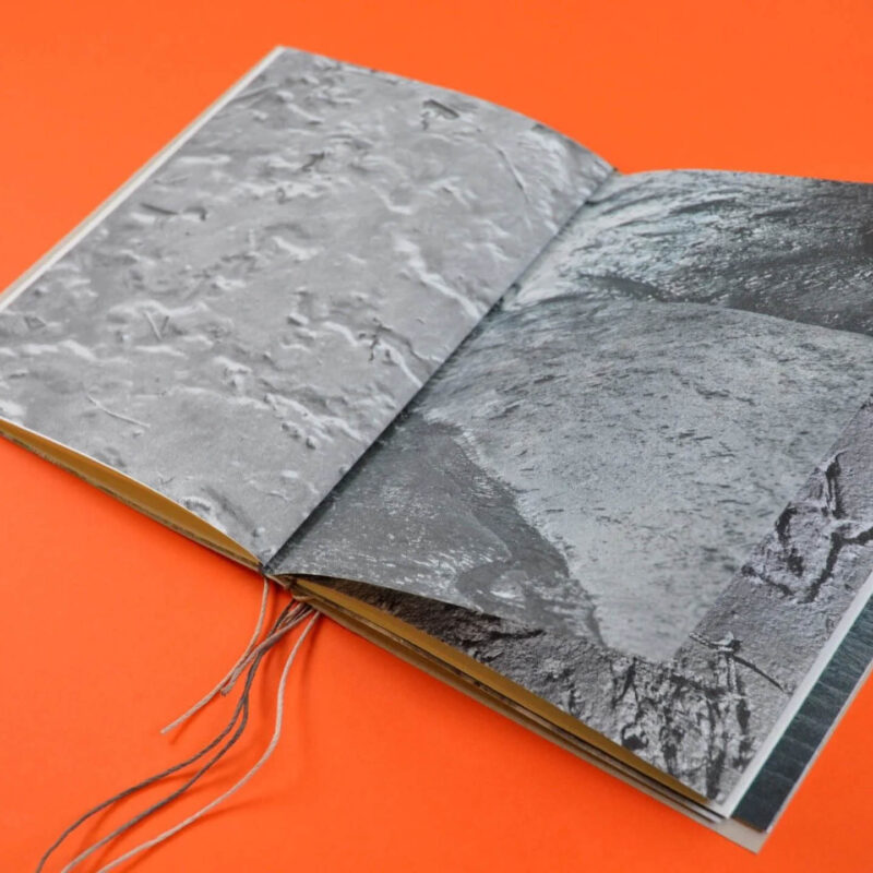 A small book lies open against an orange surface. It's pages are covered in a textured grey print. It is one of the zines on display at BALTIC for International Zine Month.