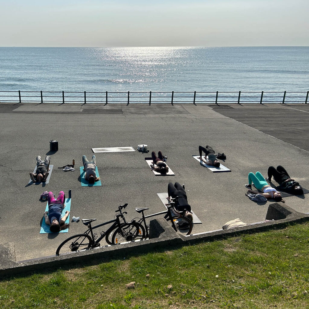 A group of people on yoga mats are lying down or stretching on a concrete promenade. Beyond the railing, the calm sea stretches to the horizon.