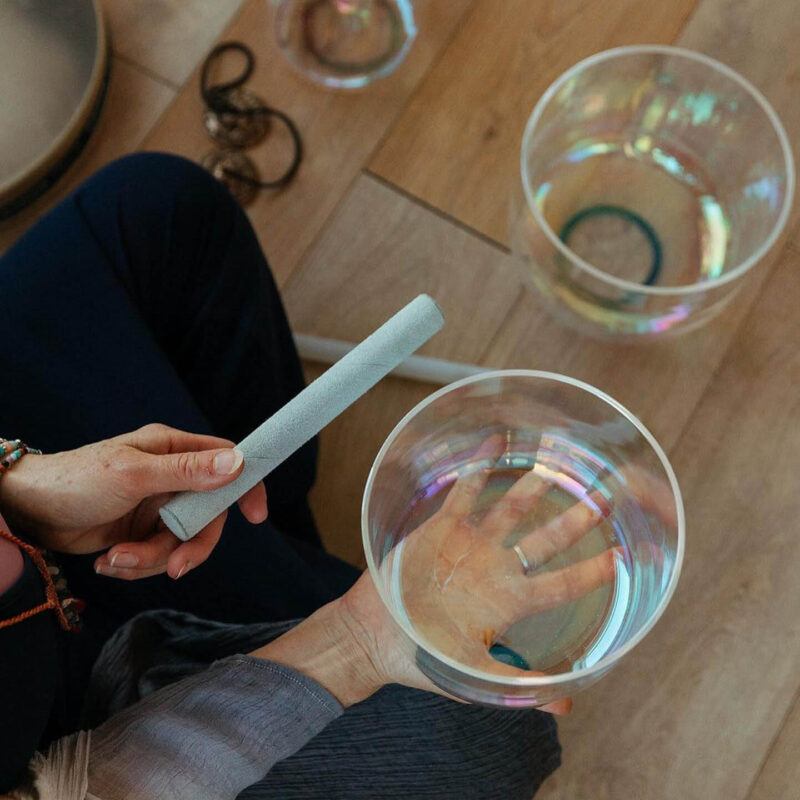 An unseen person balances a clear crystal bowl on top of their palm. They hold a small grey stick in the other hand, ready to chime it against the side of the bowl. This is part of the sound bath meditation at Jasmine Yoga in Tynemouth.
