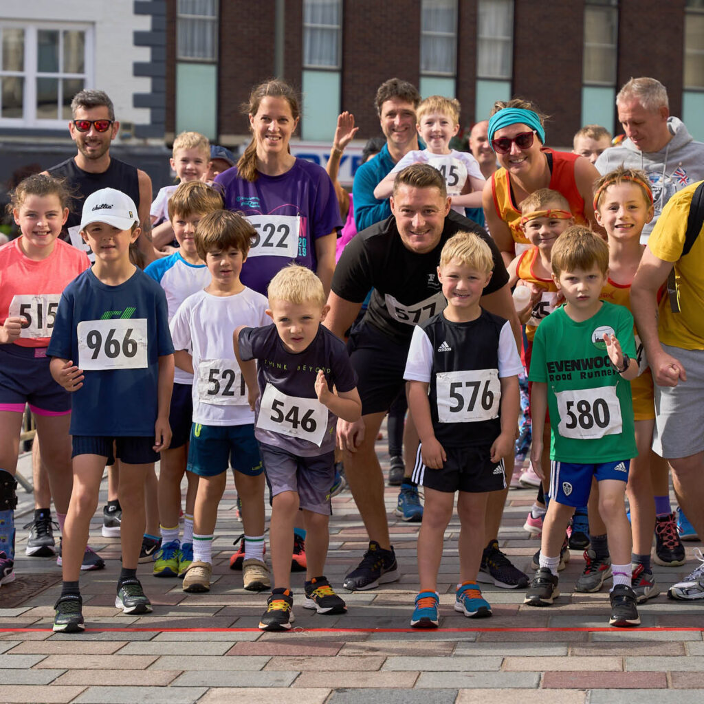 A cluster of children and adults stand at a start line, poised to start running. They all wear numbers on their t shirts and smile at the camera determinedly.