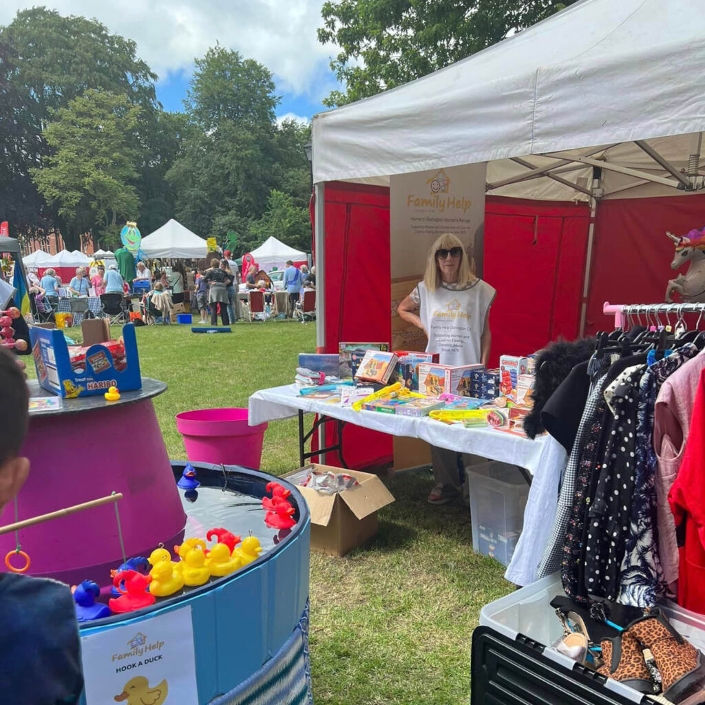 A woman stands behind her stall at Darlington Community Carnival. She is selling a range of games, puzzles and clothes. A hook a duck pool stands in the foreground. Behind the stall, other tents and crowds of people can be seen.