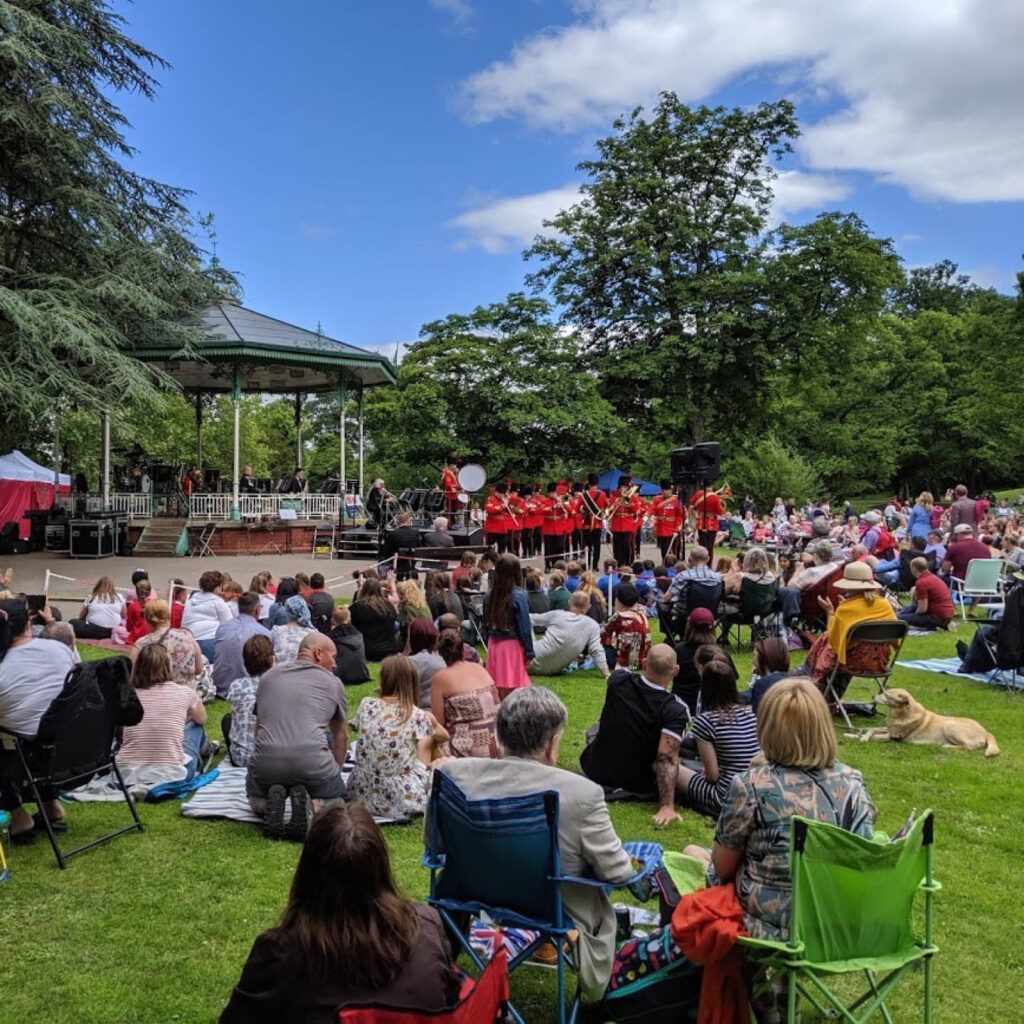 A marching brass band dressed in smart red outfits play next to the band stand in Darlington's South Park. Crowds of people watch them, most of them sitting on the grass or in their own deckchairs.