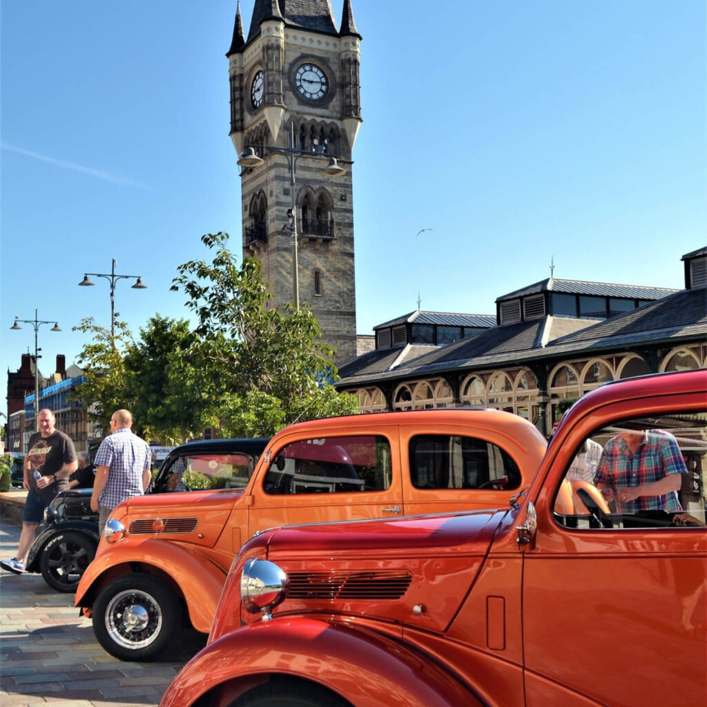 Two red vintage cars are parked beside each other in Darlington's market square. A large clock and bell tower looms over them in the background against a bright blue sky. Two men sit beside a black vintage car further along, chatting to each other.
