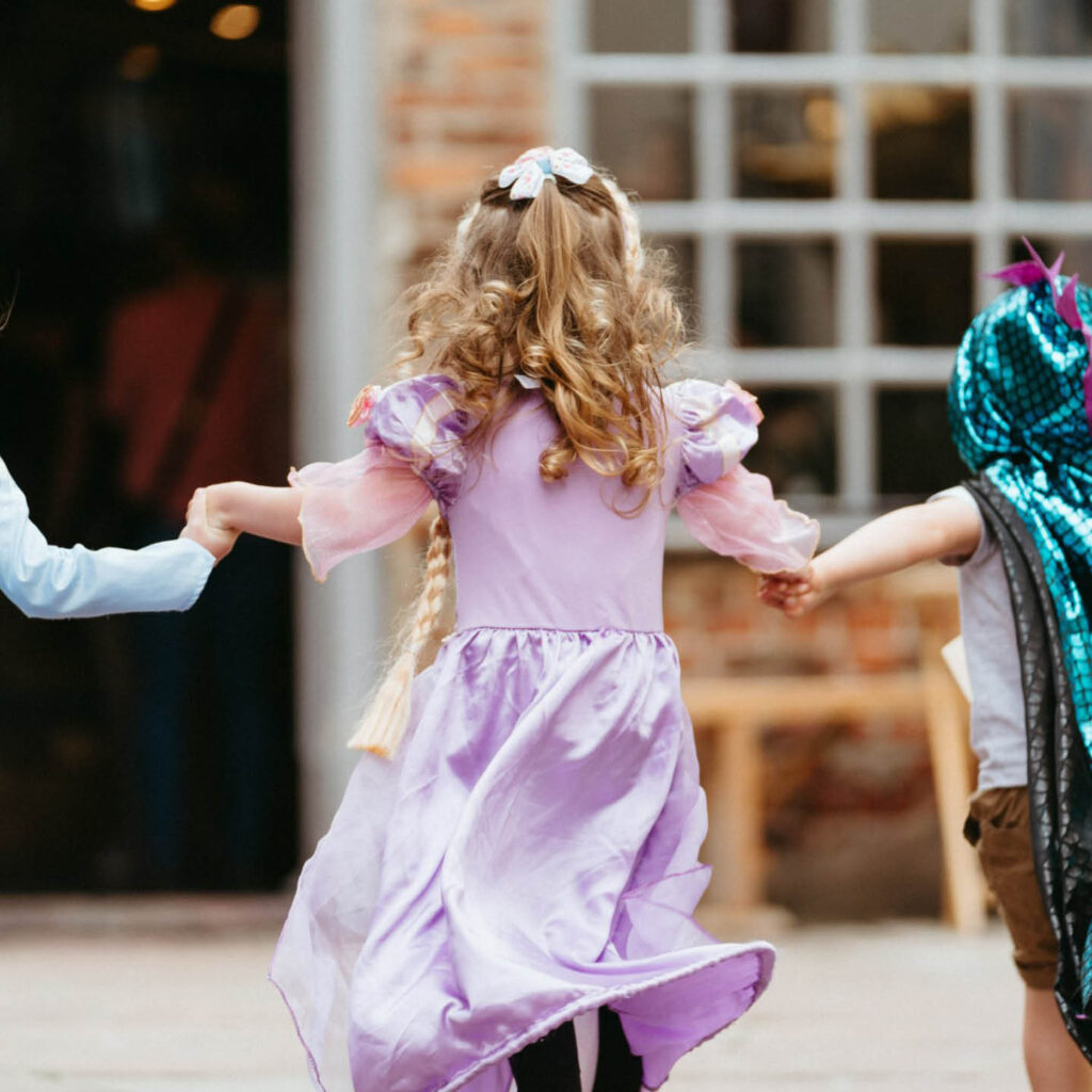 A little girl with blond ringlets runs away from the camera. She wears a pink princess dress. She holds hands with two other children, one of whom can just be seen wearing a blue iridescent dragon outfit.
