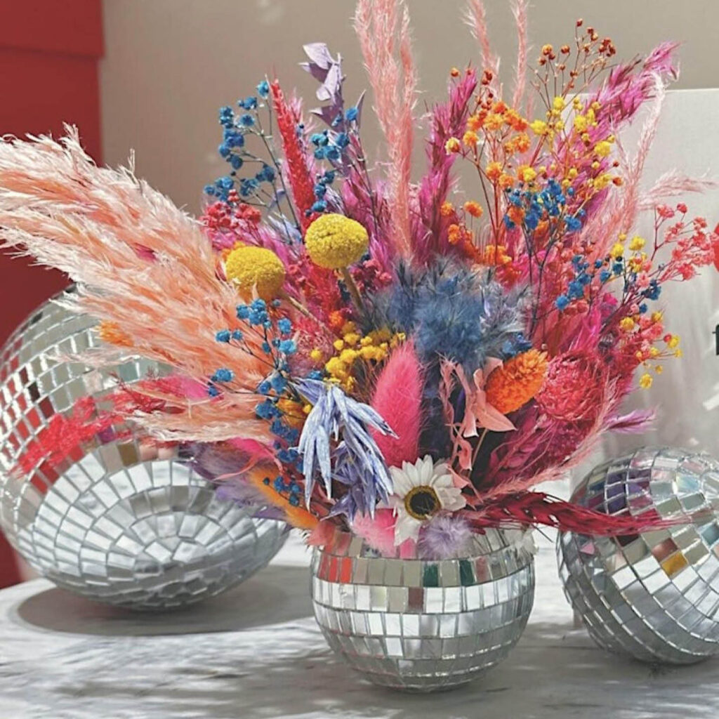 A disco ball sits on a marble surface next to others, one big and one small. A spray of vibrant dried flowers are displayed inside it.