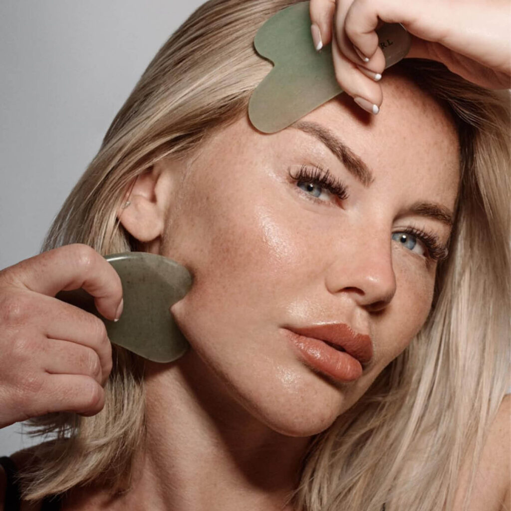 A blonde white woman with glowy skins looks at the camera, her lips pouted slightly. She drags two green gua sha stones across her temple and cheekbones.