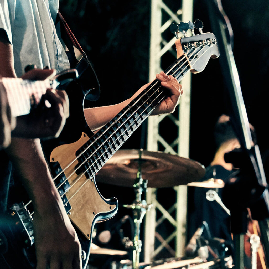An unseen musician plays an electric guitar on stage. Another musician and a drum kit stand in the background.