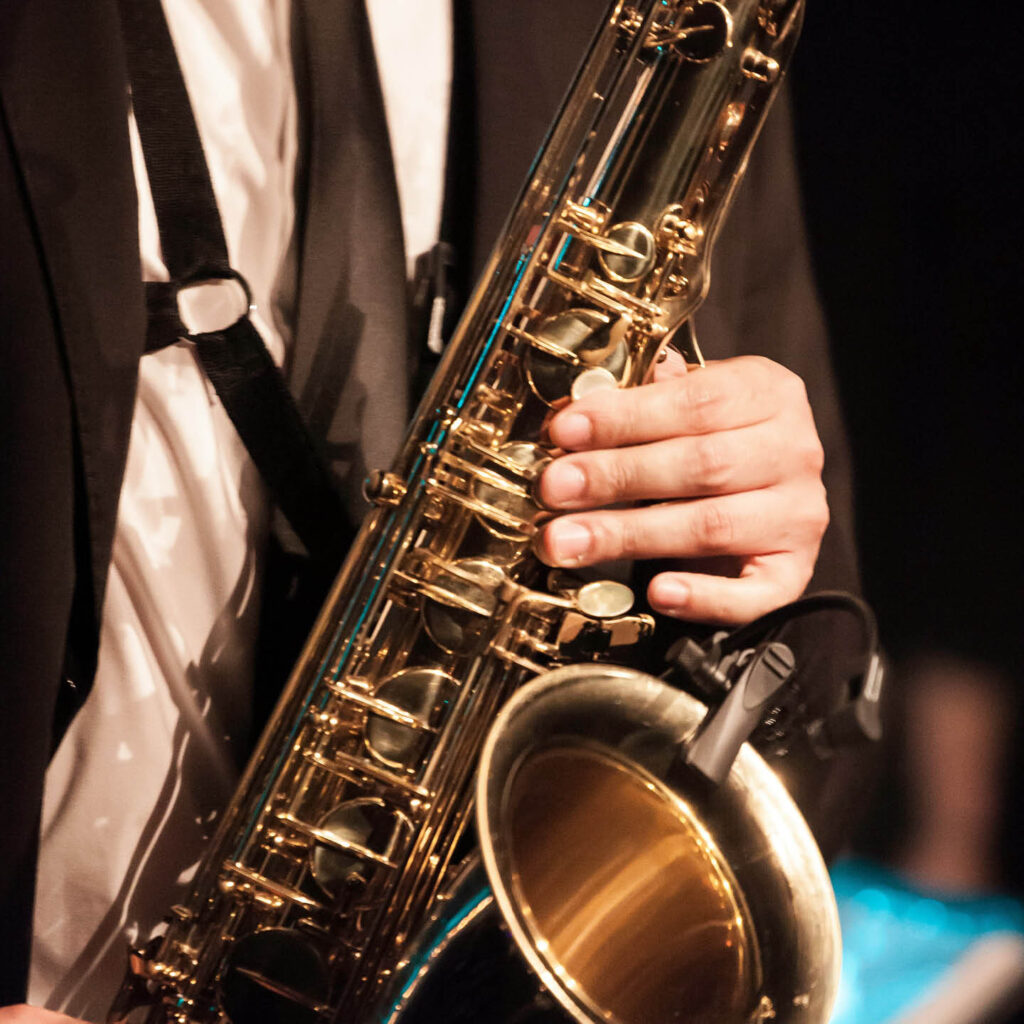 An unseen person in a suit presses their fingers against the keys of a shiny saxophone.