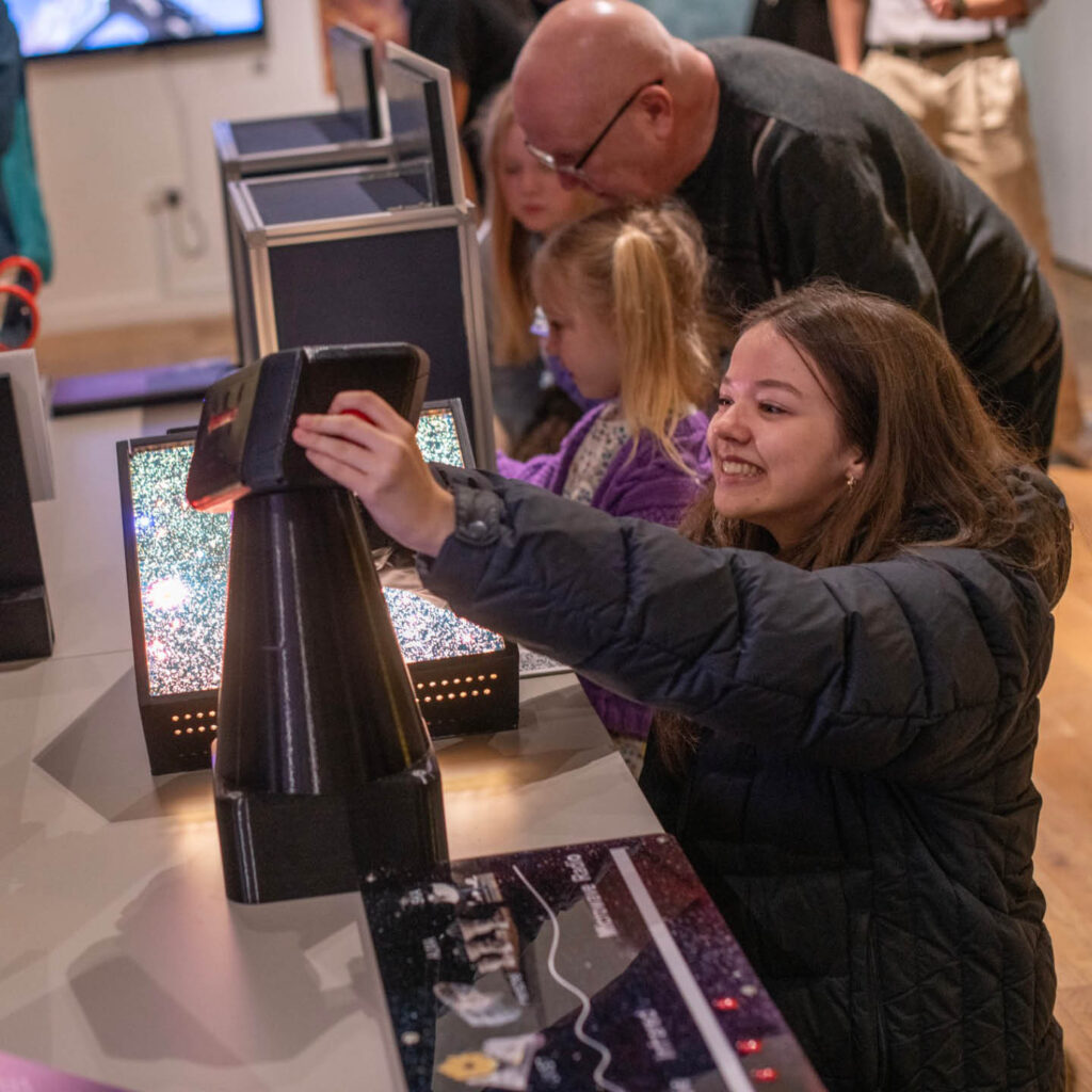 A young girl interacts with one of the exhibits at the Space Investigators exhibition at the Hancock Museum. She tilts a screen towards her and grins into it while a fuzzy screen illuminates behind her.