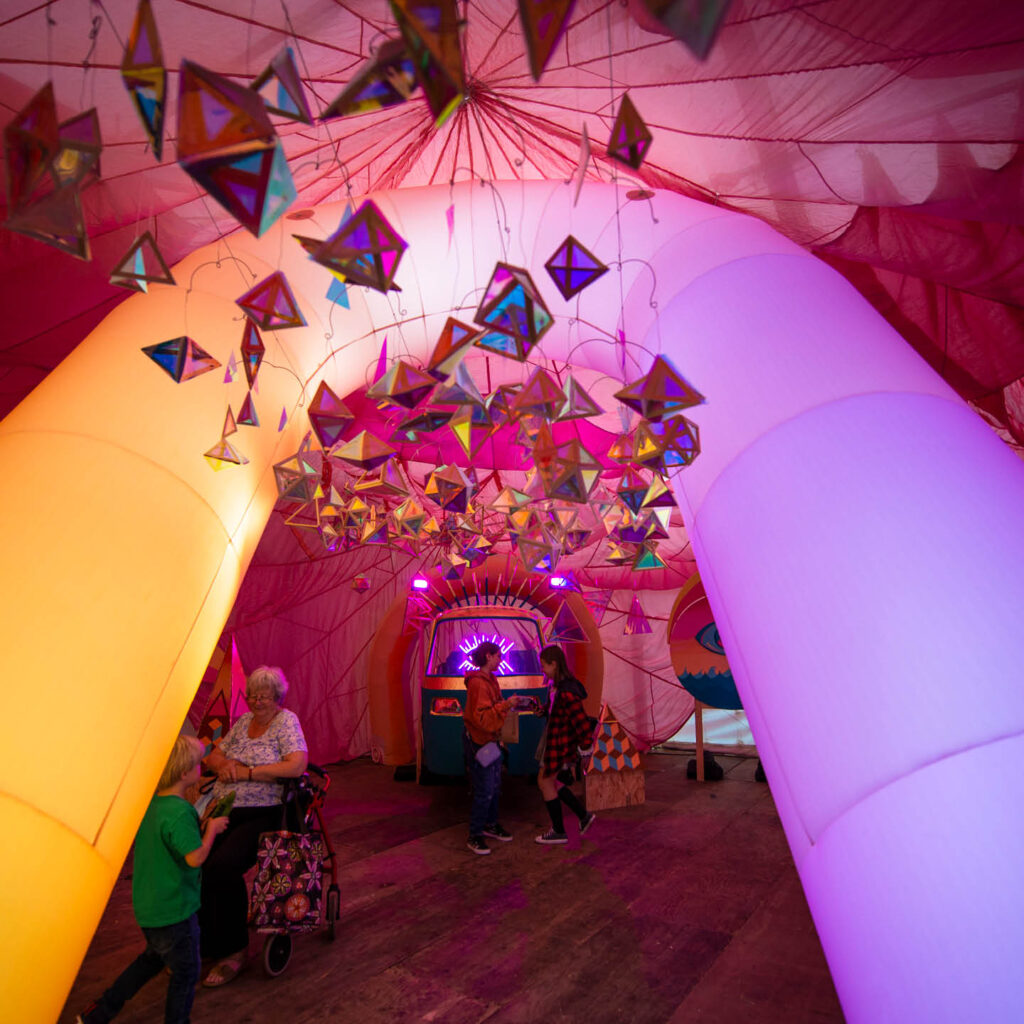 One of the experience rooms at the Happy Rooms, Middlesbrough. A spacious pink tent looms overhead, held up by a glowing yellow and purple art installation. Origami style shapes with stained glass colouring hang from the ceiling and catch the light prettily.