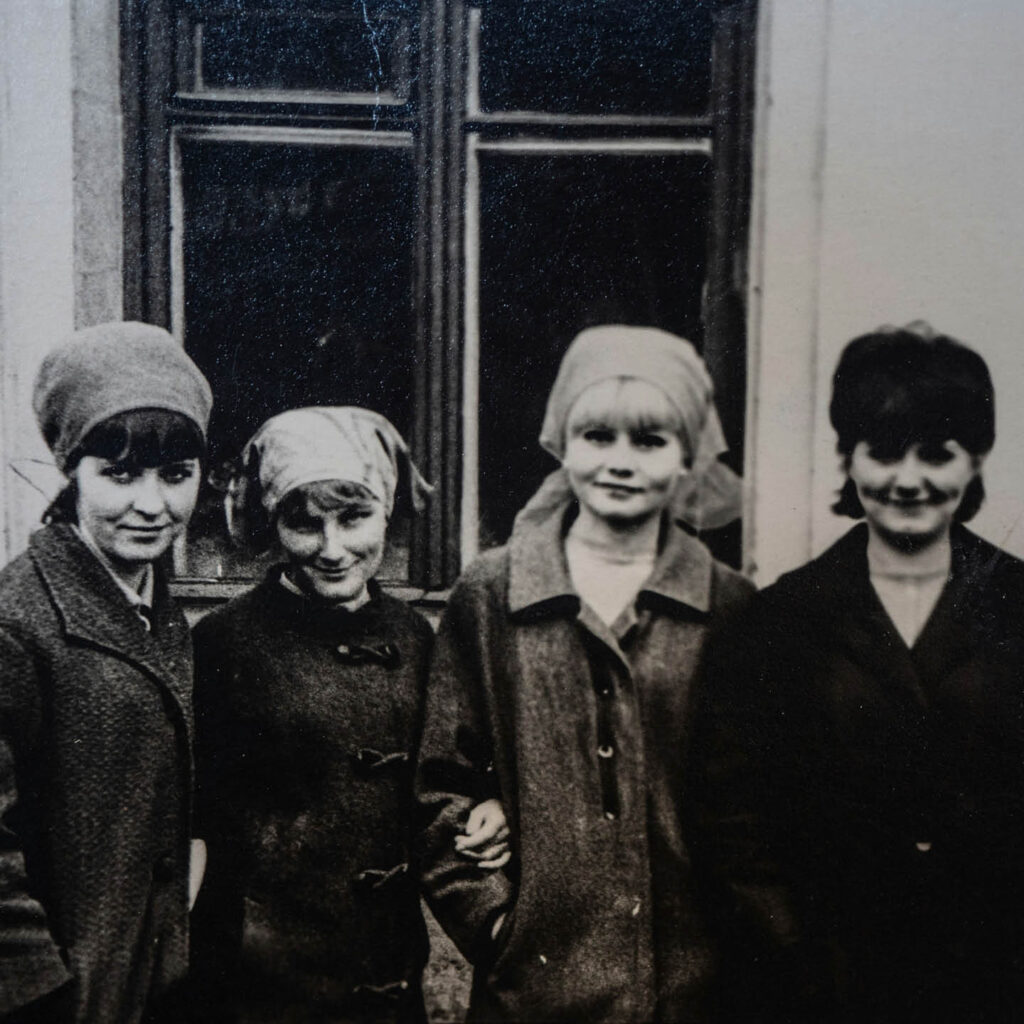 Black and white photo of four young women huddling together. Three of them wear headscarves and they all wear warm coats. They smile at the camera.