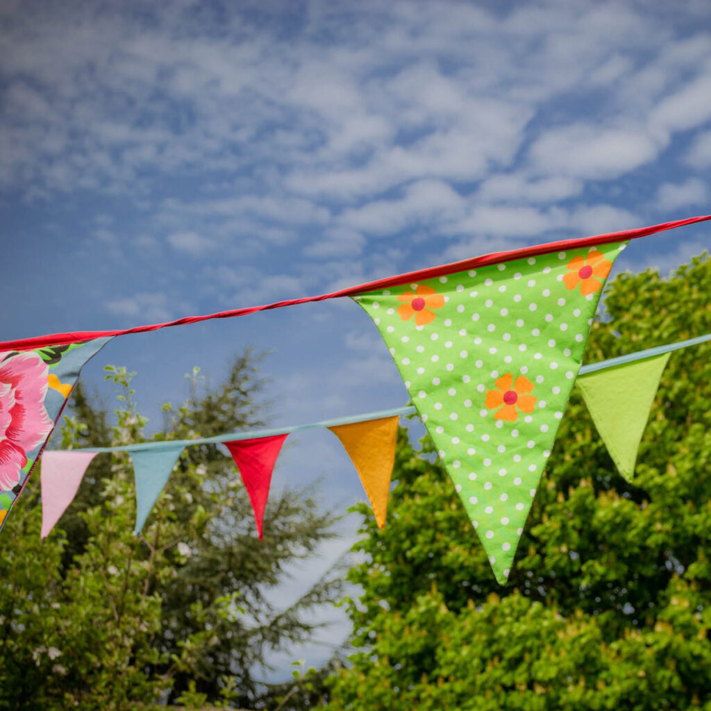 Two strings of colourful bunting hand against a bright blue sky and green trees.