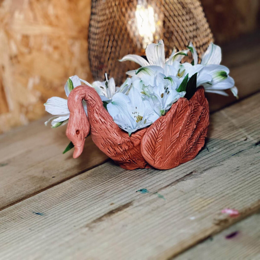A handmade terracotta swan has been made into a small vase. It holds a bunch of white flowers in its back. Its neck and wings have been carefully marked with lines for feathers.