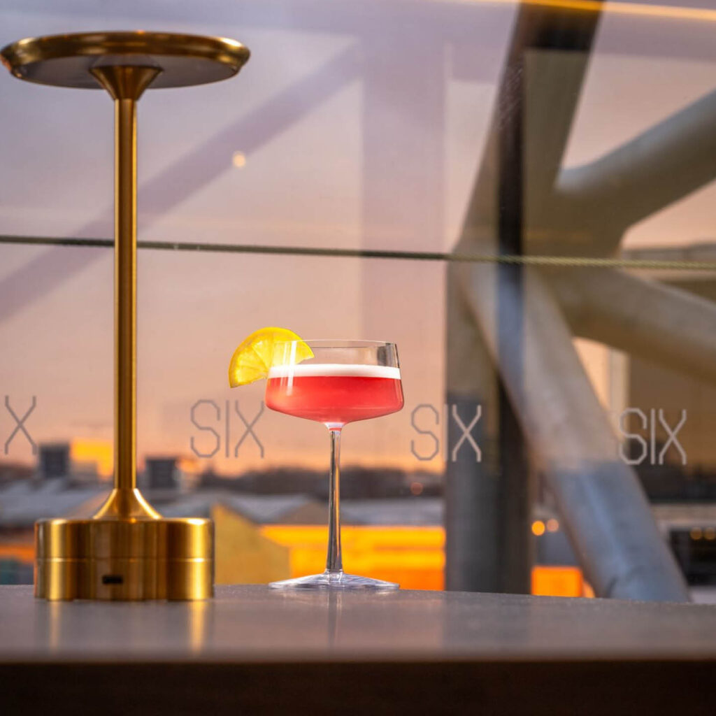 A red cocktail in a martini glass sits on a table next to a sleek gold lamp. Behind it is a large window printed with the SIX Baltic logo.