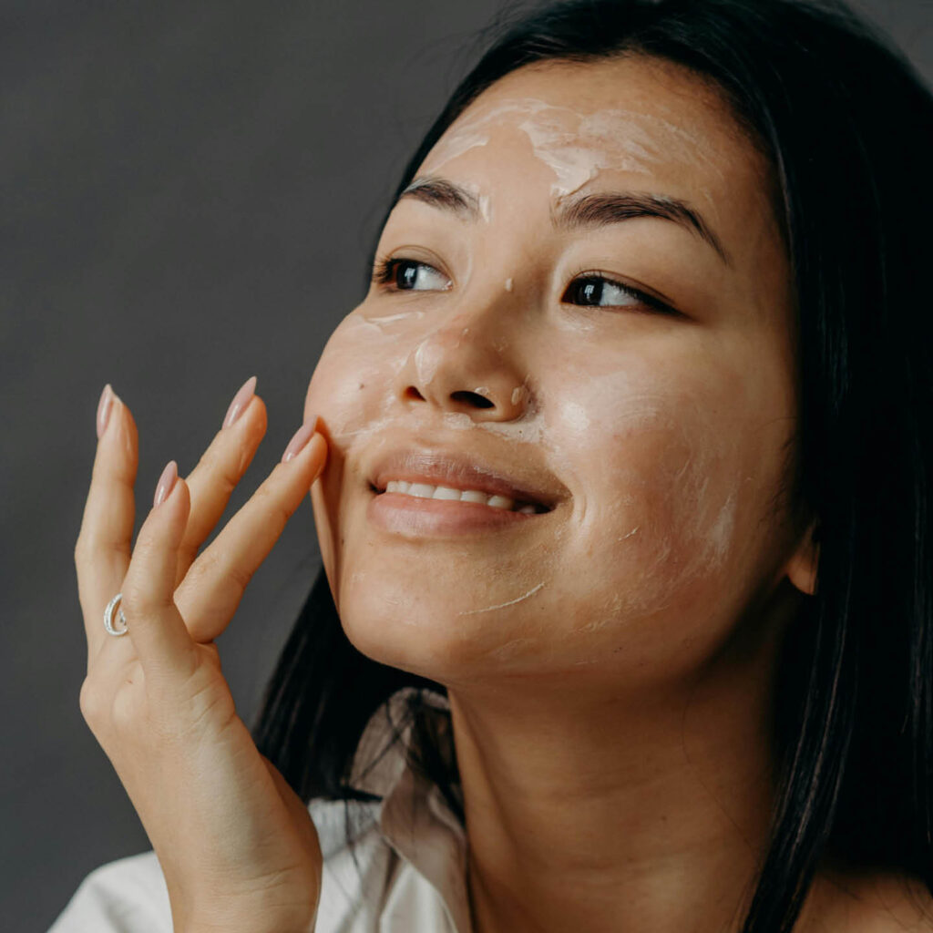 A woman smiles as she massages face cream into her skin.