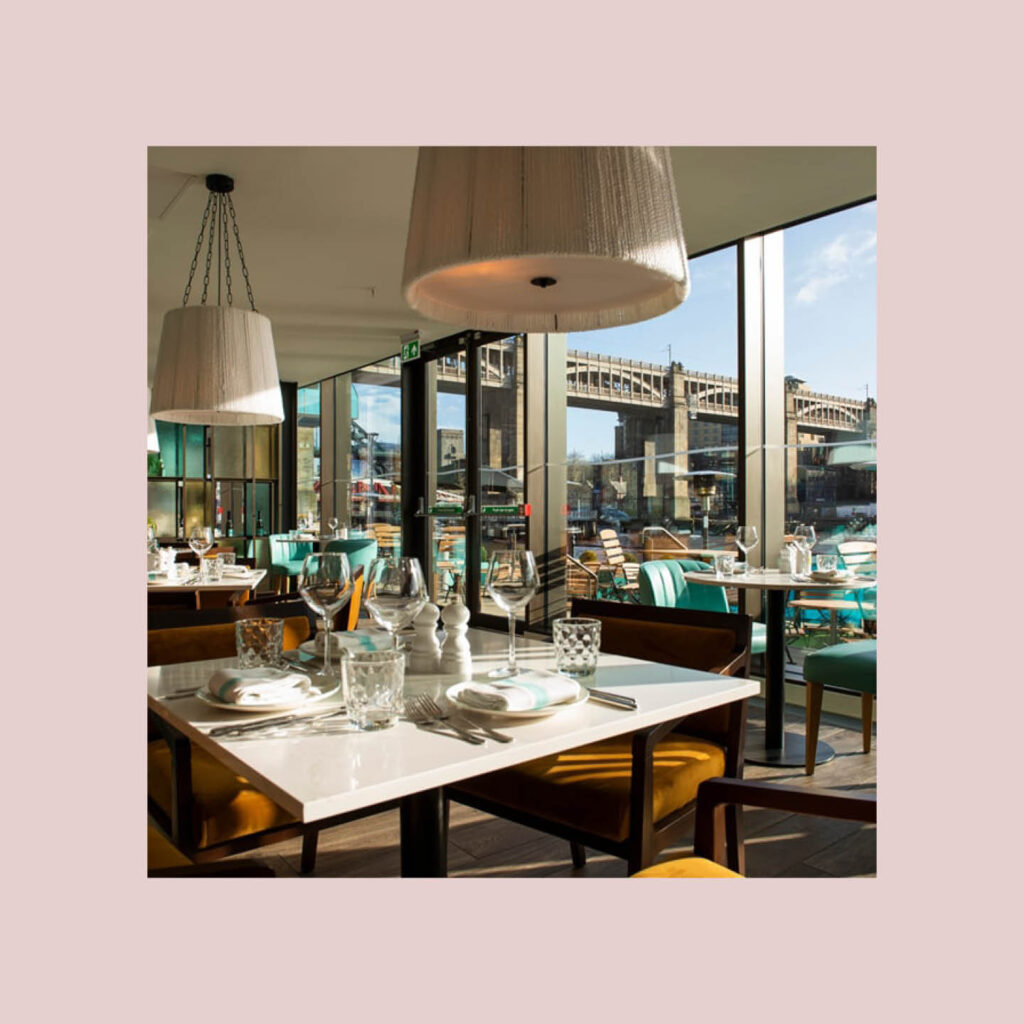 Tables are laid within the Gino d'acampo quayside restaurant in Newcastle. Sunlight fills the dining area through floor to ceiling windows, out of which one of Newcastle's bridges can be seen.