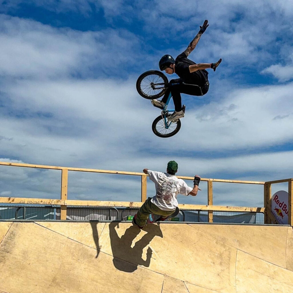 A person on a BMX hovers mid air as they come off a ramp. They are silhouetted against the blue sky. Below them, a person on a skateboard stretches out their arms as they soar up a ramp.