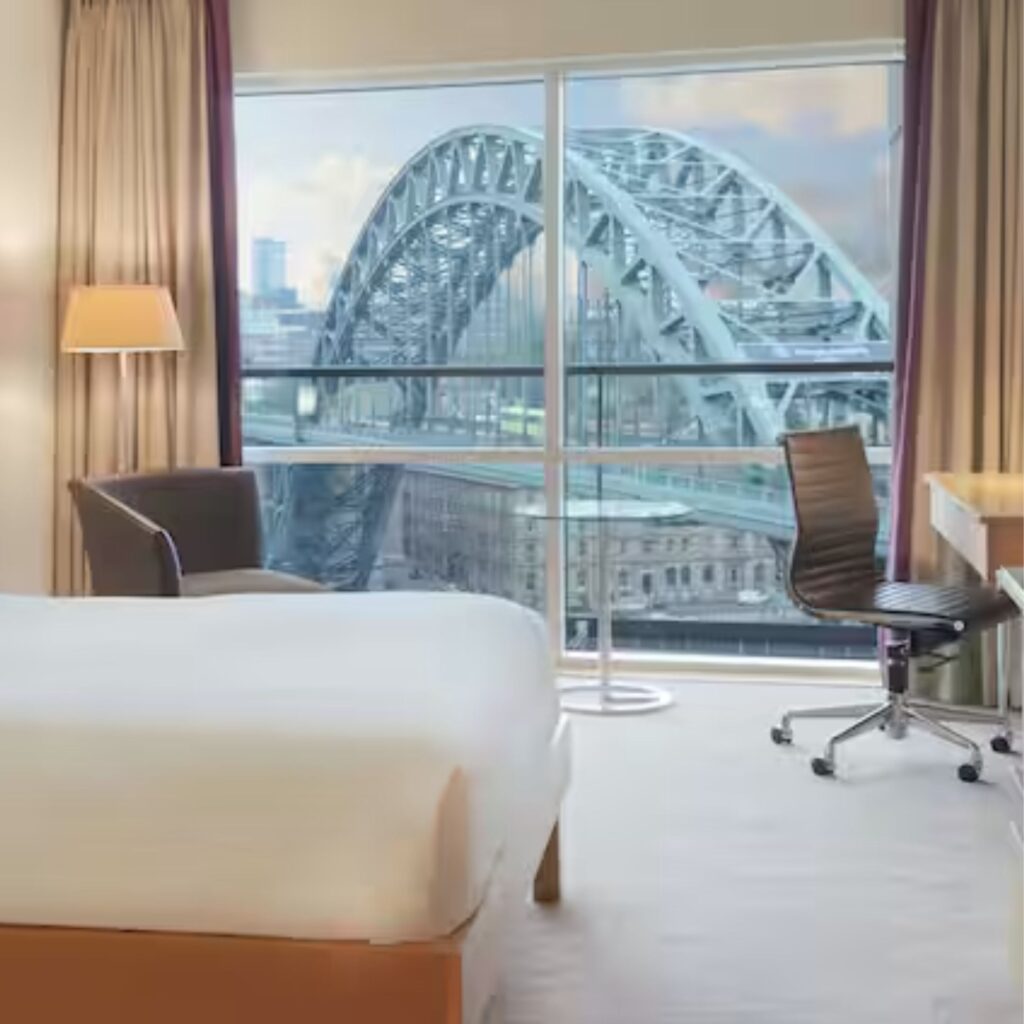 The view of the tyne bridge in Newcastle is visible from outside the window of a clean hotel room. There is a bed, desk, chair, lamp and reading corner
