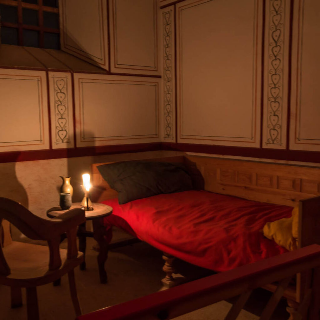 An old bedroom at Arbeia Roman Fort in South Shields. It has a small bed covered in a red sheet, an old wooden chair and a single candle lamp standing on a wooden table.