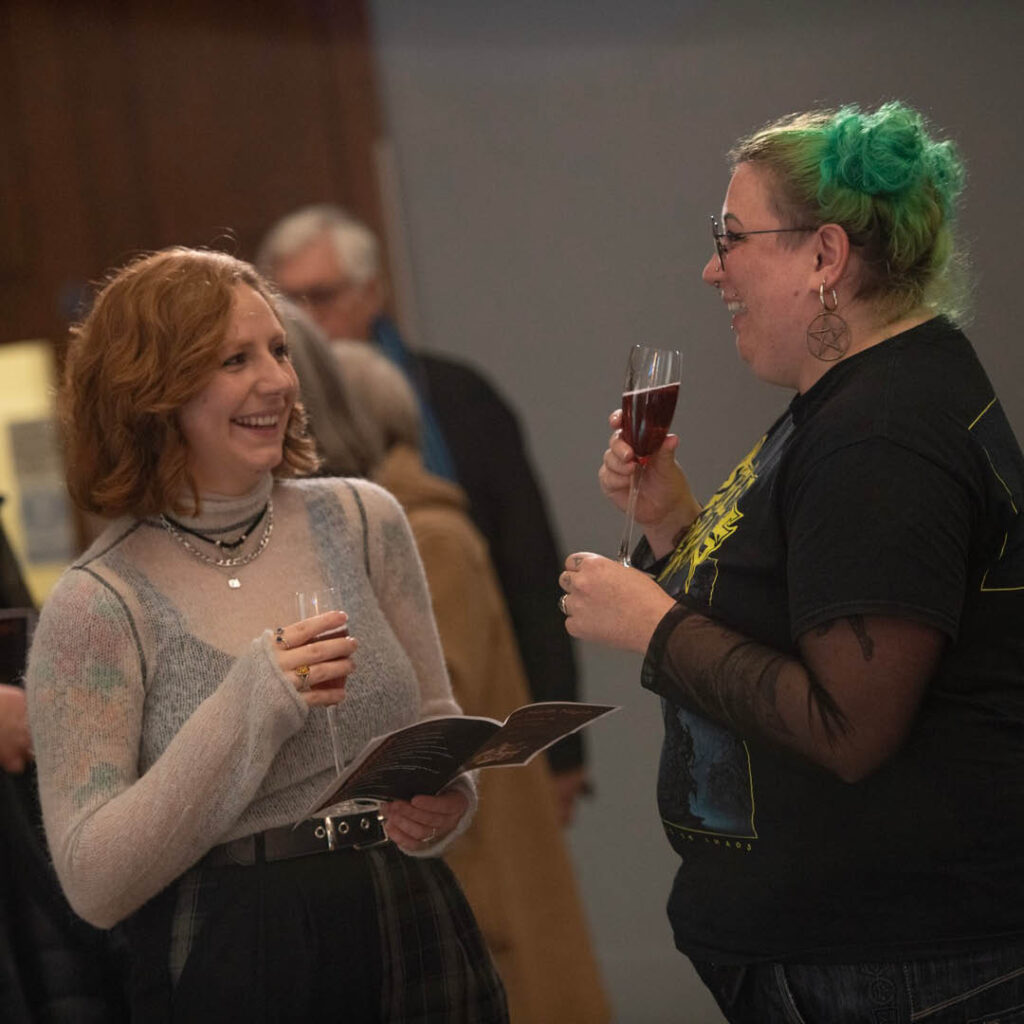 Two women laugh as they hold champagne glasses containing a red cocktail. One of them holds an open leaflet. They are at one of the Great North Hancock Museum's Great North Nights.