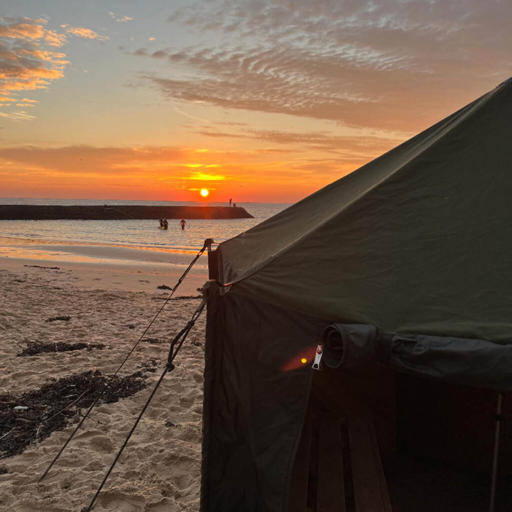 Silhouettes of people stand in the sea at Cullercoats Bay as the sun sets. In the foreground there is a dark green sauna tent.