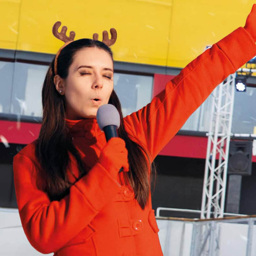 A woman wearing reindeer antlers and a bright red jacket stands on the ice rink outside of Life Science Centre. She closes her eyes as she sings into a microphone.