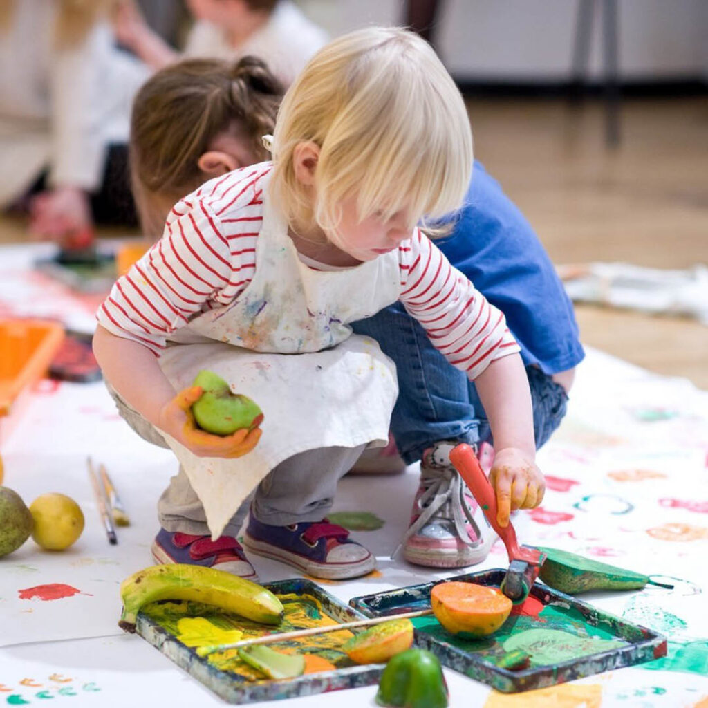 A very young child with blonde hair crouches down and dips a brush into some green paint. They hold half an apple in the other hand, ready to do some printing with it. A large white sheet of paper covers the floor beneath the child and is already covered in paint.