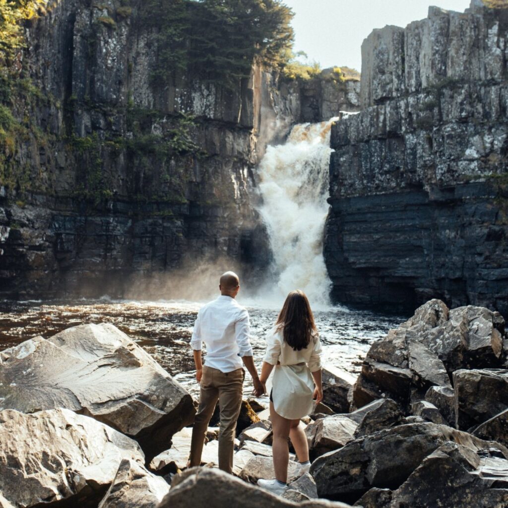 A couple hold hands as they stand on the rocks facing a large rushing water on a nice sunny day.