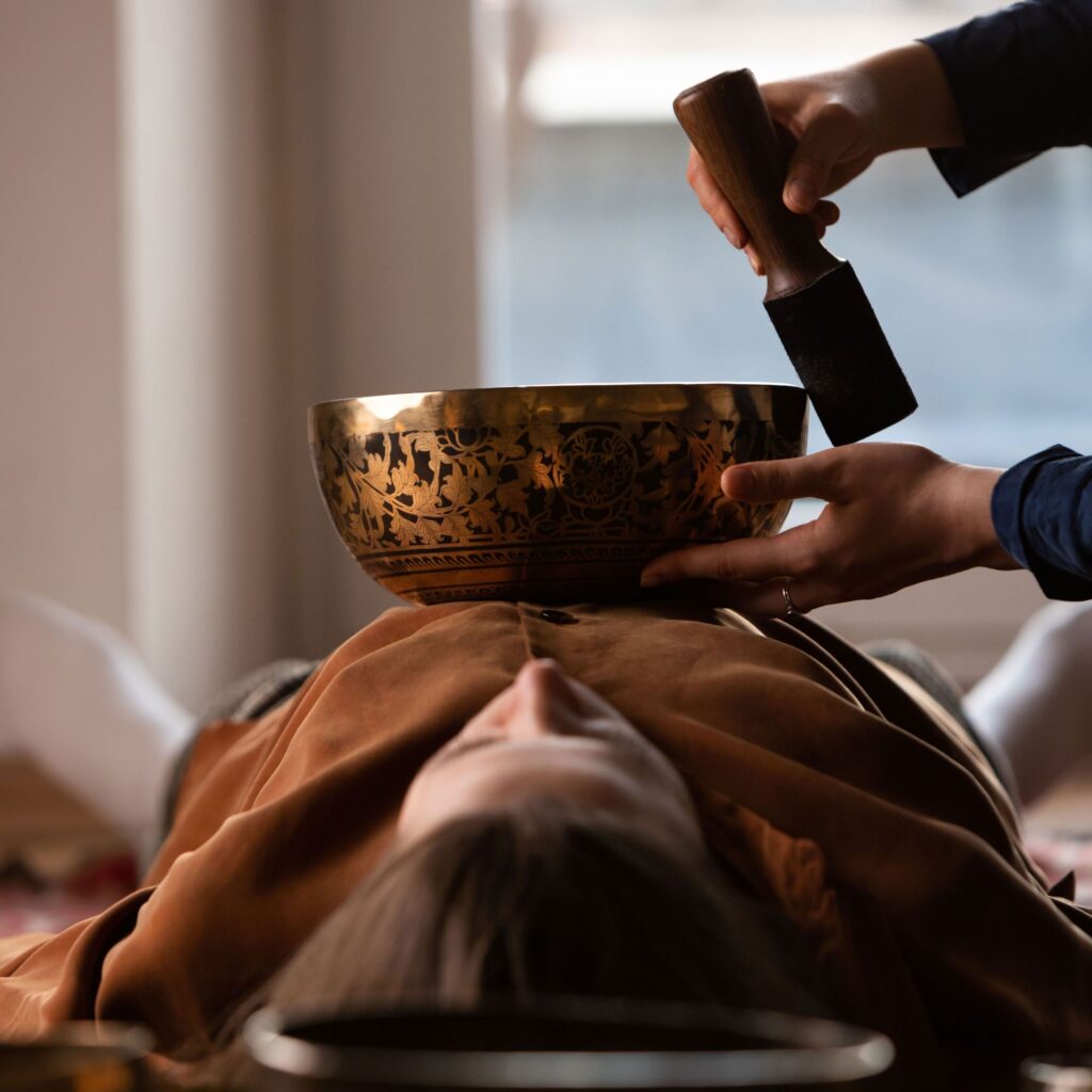A woman lays on her back in comfortable clothing while someone holds a sound bowl above her stomach