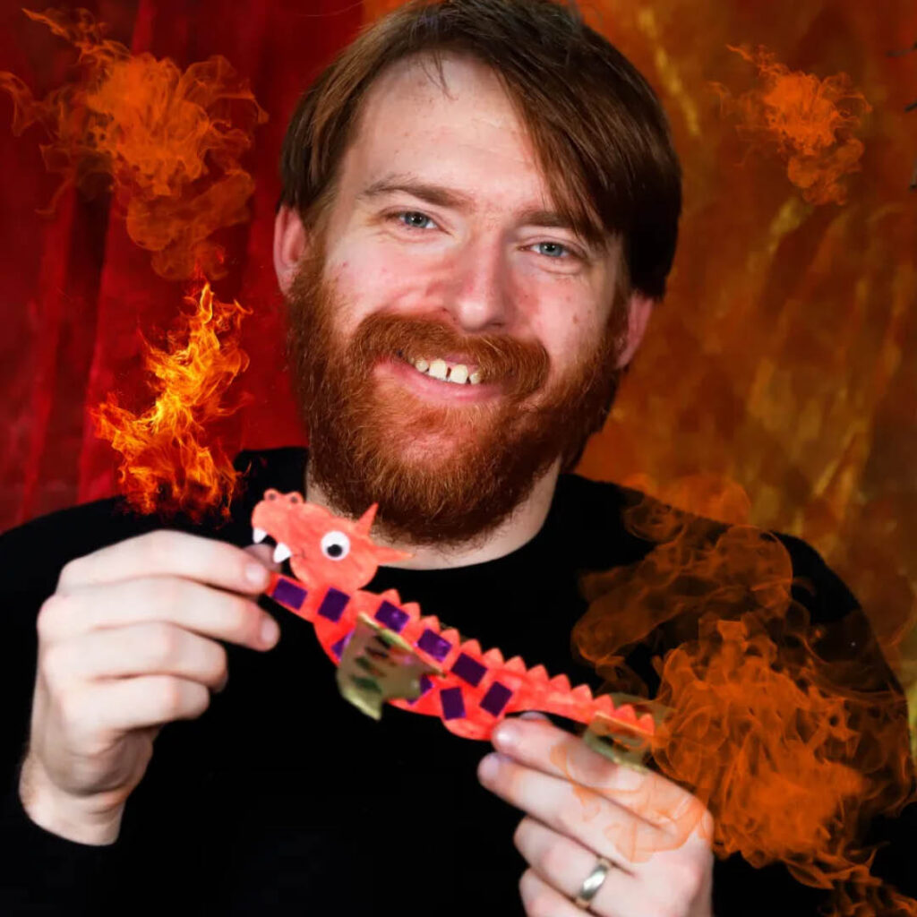 A bearded man smiles at the camera as he holds up a small red dragon with gold wings.