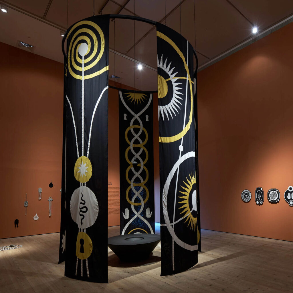 Large black banners featuring white and yellow totemic symbols hang from the ceiling to the floor in one of the Baltic's large exhibition spaces. This is part of the Mani Kambo exhibition.