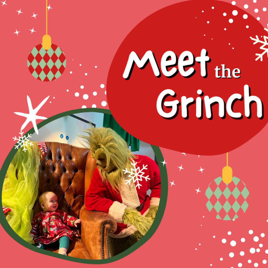 A toddler in a red Christmas top sits on a leather armchair. They smile up at the Grinch, who leans an arm on the chair.