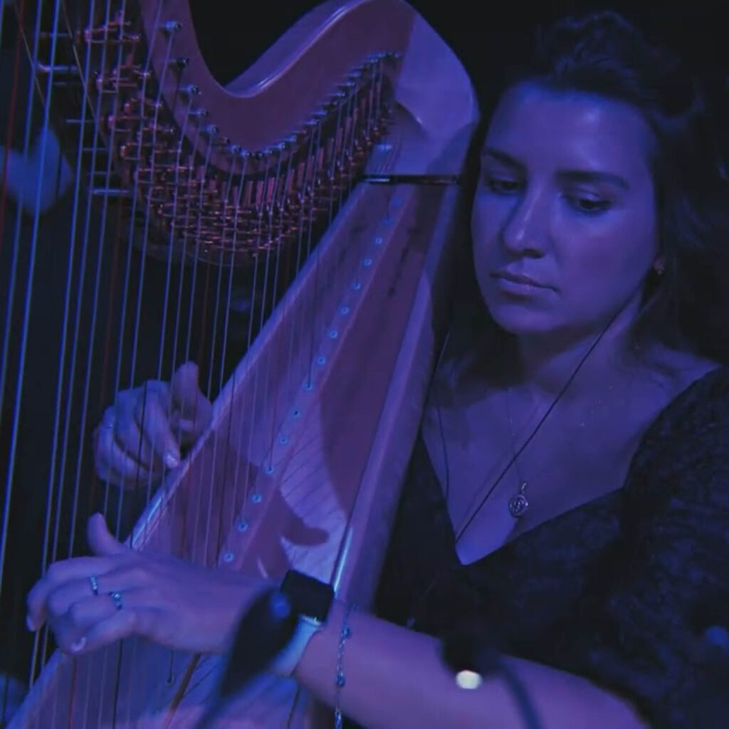 A woman plays a harp. She is cast in a deep blue and purple light. She is playing at The Glasshouse's Sci Fi concert.