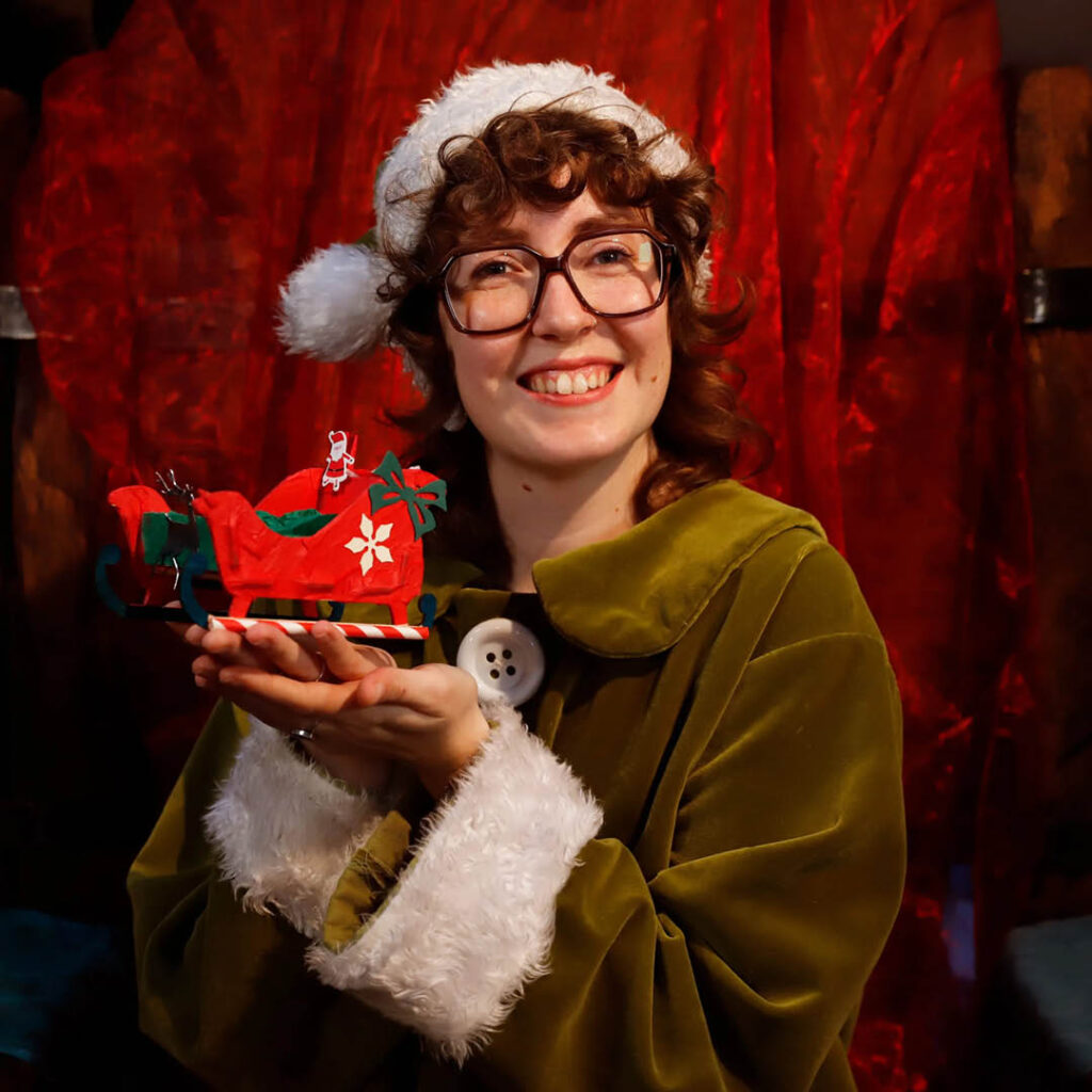 A woman in a big green coats with white fluffy cuffs and a Santa hat smiles as she holds up a mini sleigh. It is red and decorated with a snowflake and candy cane runners.