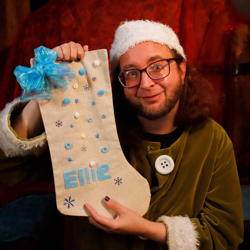 A man dressed as an elf holds up a fabric stocking embroidered with the name Ellie. It is covered in blue and white pom poms.