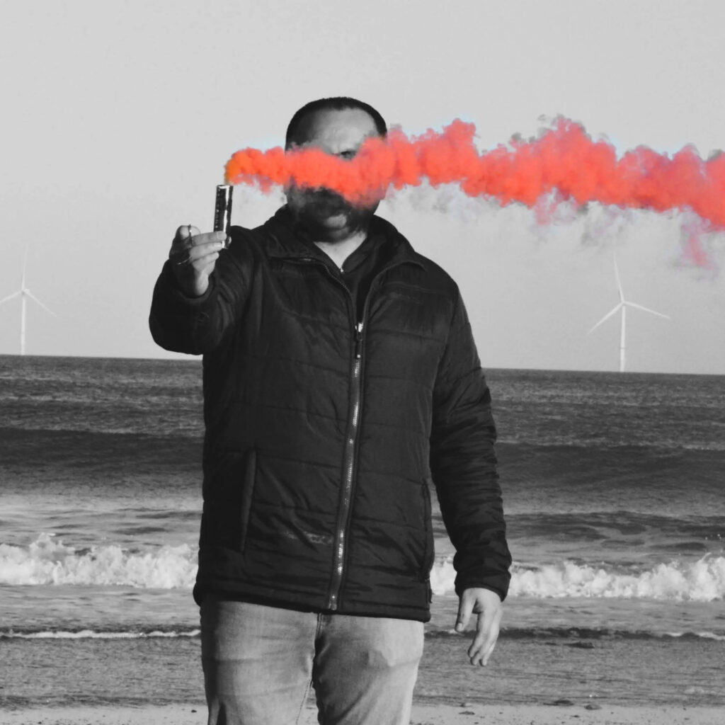 A man in a black puffer jacket stands on a beach with the waves crashing behind him. He holds up a smoke flare in front of his face. The image is black and white apart from the smoke, which is a vivid red.
